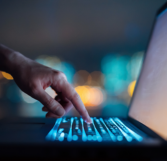 Close up of woman's hand typing on computer keyboard in the dark against colorful bokeh in background, working late on laptop at home. (source: Getty Images)