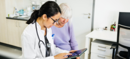 A young female doctor in a lab coat and glasses sits beside a senior patient in a modern medical office, reviewing medical information on a digital tablet. The doctor points to the screen, indicating a collaborative approach to discussing medical details or treatment plans. (source: Getty Images)